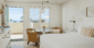 A bright, coastal-inspired room with a white bed, rattan chairs, and a round glass table, featuring an open door leading to a private balcony with ocean views